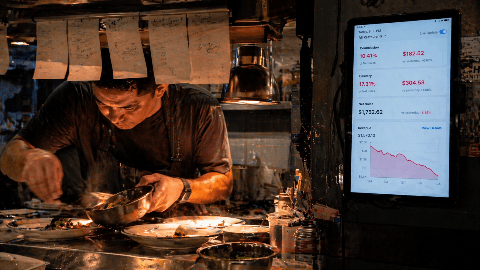 Restaurant kitchen during dinner rush - tickets on the rail, a delivery app tablet showing commission fees