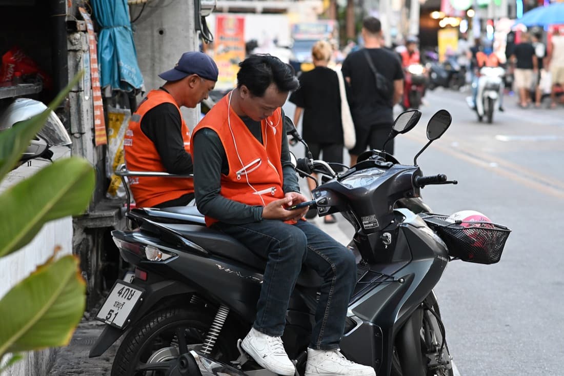 Two delivery couriers in orange vests wait on a parked scooter beside a busy street, one checking his phone