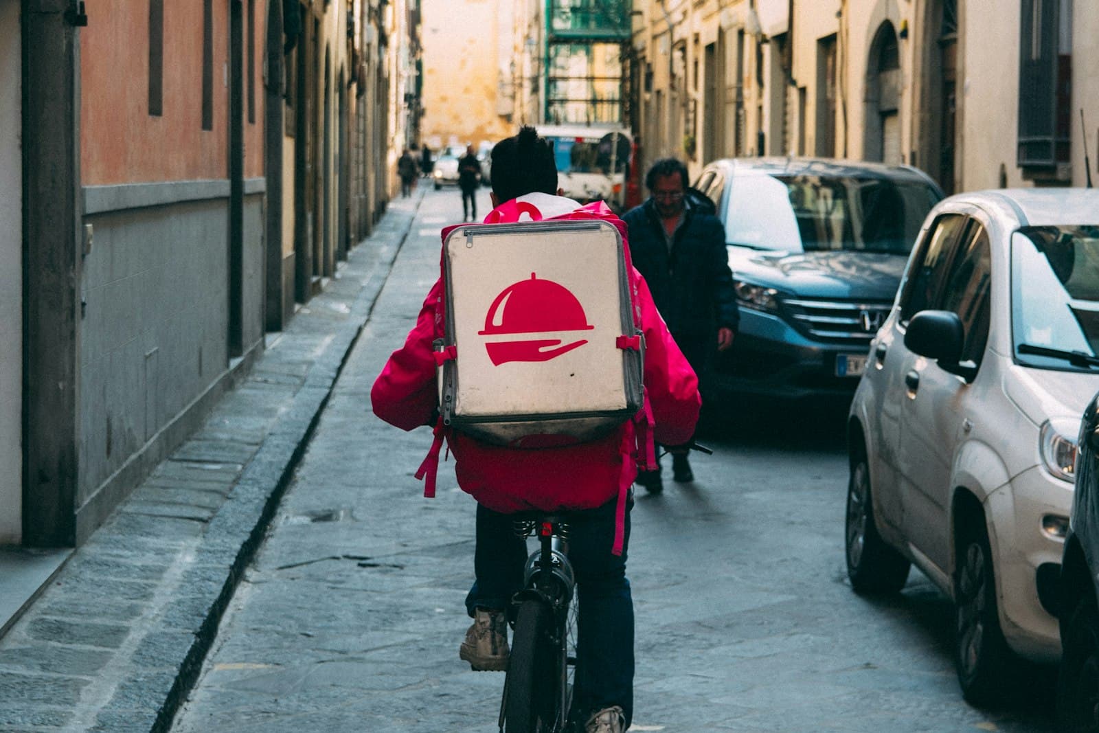 Bicycle courier in a pink jacket with an insulated delivery box riding down a narrow city street